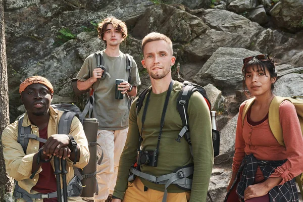 stock image Group of confident young multi-ethnic hikers with bags standing against rocks and looking at camera