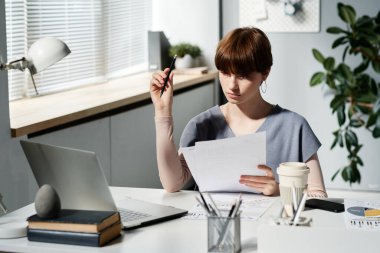 Kısa saçlı, kalem tutan ve modern ofiste kağıtları analiz eden düşünceli beyaz bir iş kadını.