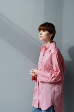 Büyük boy pembe tişörtlü düşünceli genç beyaz kadın ellerini gri arka plana yapıştırmış duruyor.