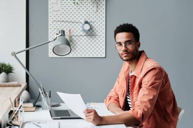 Modern ofiste elinde belgelerle oturan, gözlüklü, kendine güvenen, genç, melez bir ofis müdürünün portresi.