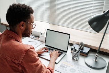 Gözlüklü genç siyahi yöneticinin omuz manzarası kapalı güneşliklerin önünde oturuyor ve dizüstü bilgisayardan bilgi arıyor.
