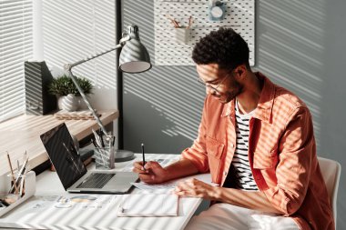 Sıradan gömlekli, konsantre, genç, melez bir işadamı masada oturuyor ve iş kâğıdına notlar yazıyor.