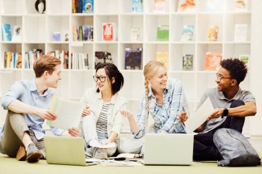 Bir grup neşeli çoklu etnik öğrenci kütüphanede yerde oturuyor ve birlikte bilgi edinirken kağıt ve dizüstü bilgisayar kullanıyorlar.