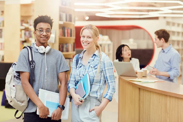 stock image Cheerful multi-ethnic teenage students in casual outfits standing in school library and holding workbooks while posing for camera