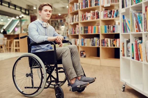 Bacakları felçli öğrenci portresi tekerlekli sandalyede oturuyor ve kütüphanede kitap seçiyor.