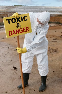 Beyaz koruyucu giysi ve maskeli ekoloji uzmanı. Kirli bölgeye zehirli tehlike işareti yerleştiriyor.
