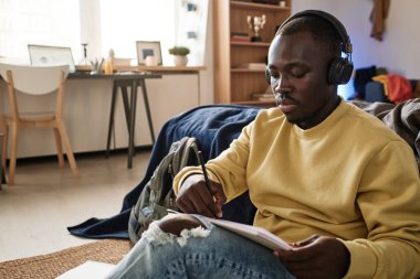 Kulaklıklı ve sarı kazaklı genç Afro-Amerikan öğrenci kulaklıkla müzik dinliyor ve çizim defterinde çizim yapıyor.