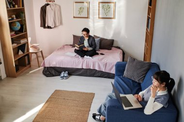 Hazırlıklı kız kampüs odasındaki dizüstü bilgisayarda makale okurken Asyalı öğrenci ders kitabı okuyor.