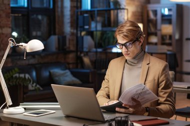 Boğazlı kazaklı genç ciddi bir işadamı laptopuyla masada oturuyor ve karanlık ofiste kağıtları analiz ediyor.