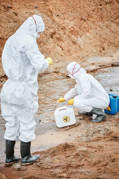 stock image Ecological researchers in hazmat suits and rubber boots taking dirty water substance with pipette while studying it on polluted territory