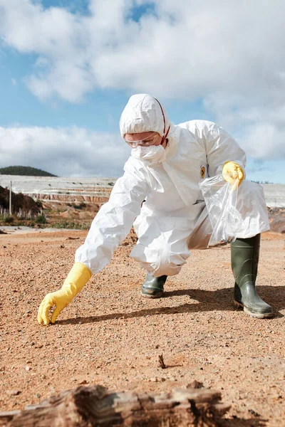 Maskeli ve güvenlik gözlüklü bir çevre araştırmacısı toprak numunesini kirlenmiş topraklarda plastik torbaya koyar.