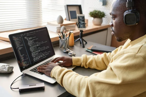 Concentrated African American app developer in yellow sweater sitting at table and working on code using laptop