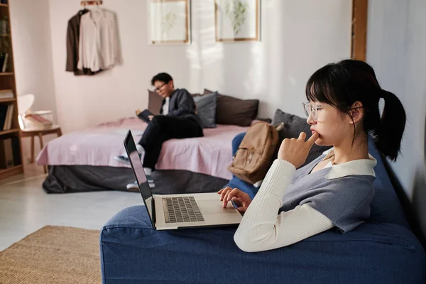 stock image Serious pensive Asian student girl in eyeglasses sitting on sofa and using laptop while browsing internet in students room