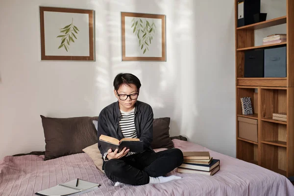 Serious young Asian college guy in glasses sitting with stack of books and reading it in bedroom