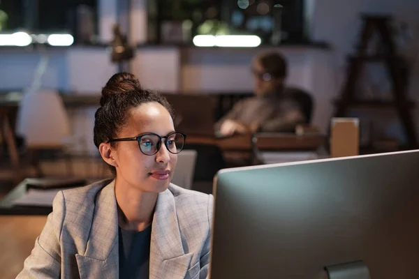 Gözlüklü ve ceketli genç kadın analist Nigth 'te ofisinde oturuyor ve çevrimiçi veri üzerinde çalışıyor.