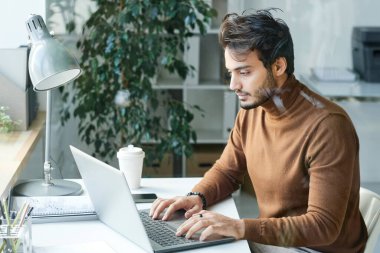 Kahverengi balıkçı yakalı konsantre genç adam masada oturuyor ve bilgisayarını ofiste kullanıyor.