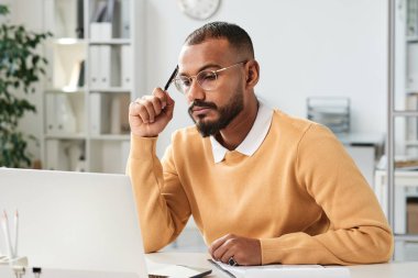 Ciddi meşgul, orta doğulu, sakallı bir pazarlamacı elinde kalemle ofiste strateji üzerinde çalışırken dizüstü bilgisayar ekranına bakıyor.