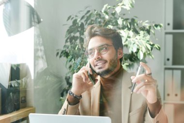 Gözlüklü neşeli, kendinden emin, genç bir ortadoğulu müdürün cam manzarasının arkasında elinde kalem, telefonda konuşuyor.