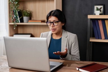 Kendine güvenen, ceketli genç İspanyolca öğretmeni ofiste oturuyor ve video konferans uygulaması üzerinden öğrenciyle konuşurken dizüstü bilgisayar kullanıyor.