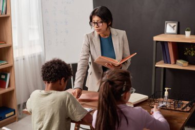 Sınıfta öğrencinin hatasını yorumlarken ceketli, ciddi bir İspanyol öğretmeni ders kitabına işaret ediyor.