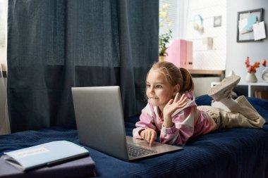 Sevimli sarı saçlı kız yatakta uzanıyor ve internette öğretmenle konuşurken el sallıyor.