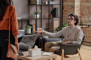 Modern genç psikoloğun kadınlara koltukta oturmayı teklif ettiği yatay bir fotoğraf.