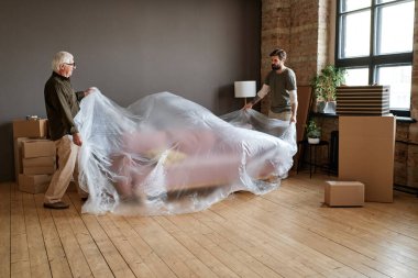 Yaşlı beyaz bir adamın, oğlunun başka bir daireye taşınmasına yardım ettiği, plastik film ile paketlenmiş bir kanepe.