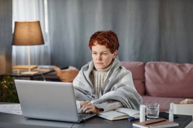 Soğuk algınlığı geçiren, kalın giysiler giyen olgun bir kadının portresi. Oturma odasında dizüstü bilgisayarla çalışıyor.