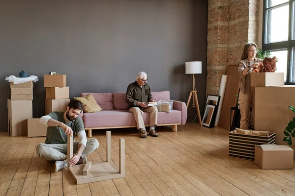 stock image Horizontal long shot of young man and his girlfriend helping grandpa to unpack stuff and assemble furniture after moving to new house