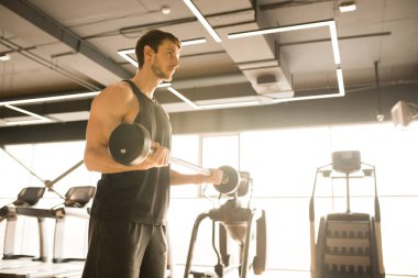 Ciddi genç adam ağırlık kaldırıyor odaklanmış görünüyor, spor salonunda tek başına çalışıyor.