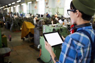 Ensesinde kulaklık olan düşünceli kadın mühendisin fabrikadaki üretim atölyesini incelerken dijital tablet kullanması.