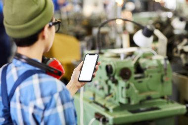 Şapkalı bayan mühendisin endüstriyel dükkanda dikiz aynasından akıllı telefon kullanarak bilgi taraması yapması.