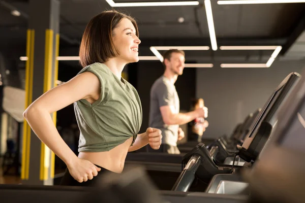 Happy young woman working out in gym doing cardio training on treadmill together with young man in the background