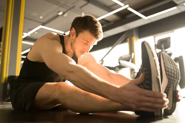 Young muscular man sitting and stretching his legs with eyes closed during sports training in gym