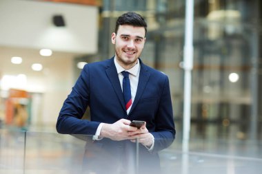 Neşeli, kendine güvenen, koyu takım elbiseli yakışıklı iş adamı giriş salonunda kameraya bakarken akıllı telefondan mesaj atıyor.