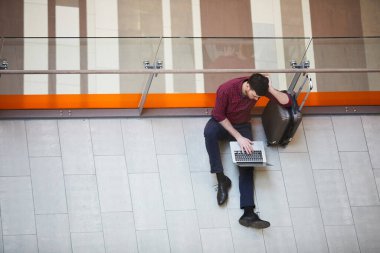 Pantolonlu, sıradan tişörtlü, yerde oturan ve havaalanı koridorundaki valizlere yaslanan ciddi bir gencin en iyi görüntüsü.