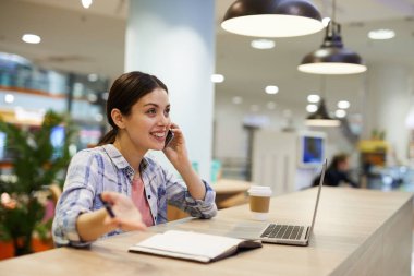 Genç, çekici, serbest çalışan bir kadın. Günlük gömlekli, kahve dükkanında oturuyor ve telefonla konuşurken el kol hareketi yapıyor.