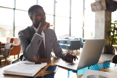 Gri takım elbiseli, düşünceli, genç bir iş adamı, ahşap masada oturmuş, internette bilgi topluyor.