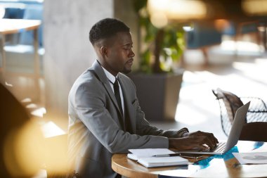 Ciddi derecede meşgul, sakallı, genç, Afro-Amerikalı iş adamı masada oturuyor ve internette gezinirken dizüstü bilgisayarında yazıyor.