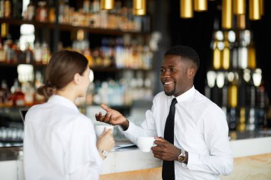 Beyaz tişörtlü, bar tezgahına yaslanmış ve molada kahve içerken sohbet eden neşeli, çok ırklı iş adamları.