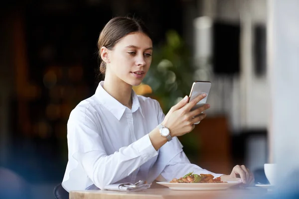 Content young business lady in white blouse sitting at table with dish and checking messenger on smartphone while having business lunch in restaurant