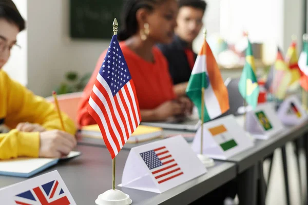 Close-up of flags of different countries standing on table with multiracial students studying in background