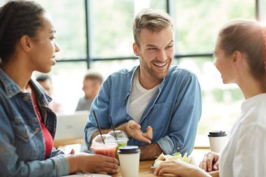 Bir grup neşeli, çok ırklı öğrenci masaya yaslanıp, sohbet ederken fast food yiyorlar.