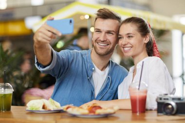 Neşeli, heyecanlı, günlük giysiler içinde genç bir çift masada oturuyor ve kafede selfie çekiyor.