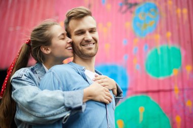 Kapalı gözlü, olumlu, sevecen genç bir çift. Duvarın önünde durmuş birbirlerine sarılıyorlar.