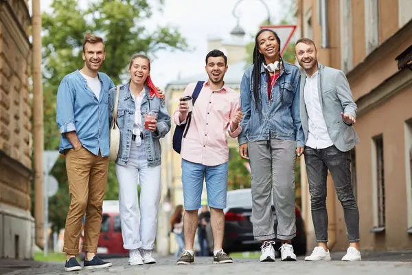 Group of excited young multiethnic friends in hipster outfits standing on street and looking at camera cheerfully