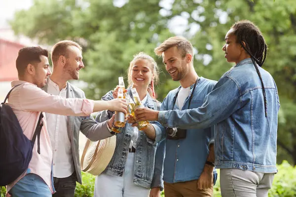 Group of cheerful young interracial people standing in city park and clinking bottles while drinking to friendship