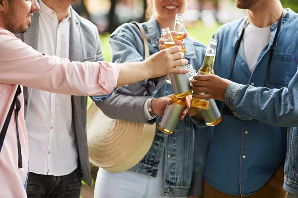 Close-up of excited young friends standing in circle and clinking bottles while drinking beer outside