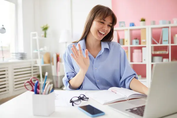 Happy excited young office lady with bangs sitting in front of laptop and waving hand while talking via video conference
