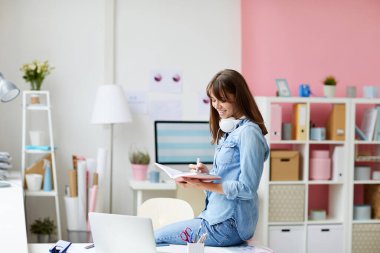 Kahverengi saçlı neşeli, yaratıcı genç kadın masaya oturmuş günlükteki notları analiz ediyor.